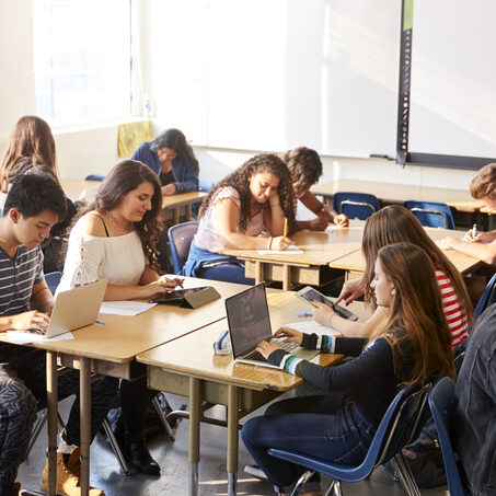 Wide Angle View Of High School Students Sitting At Desks In Classroom Using Laptops